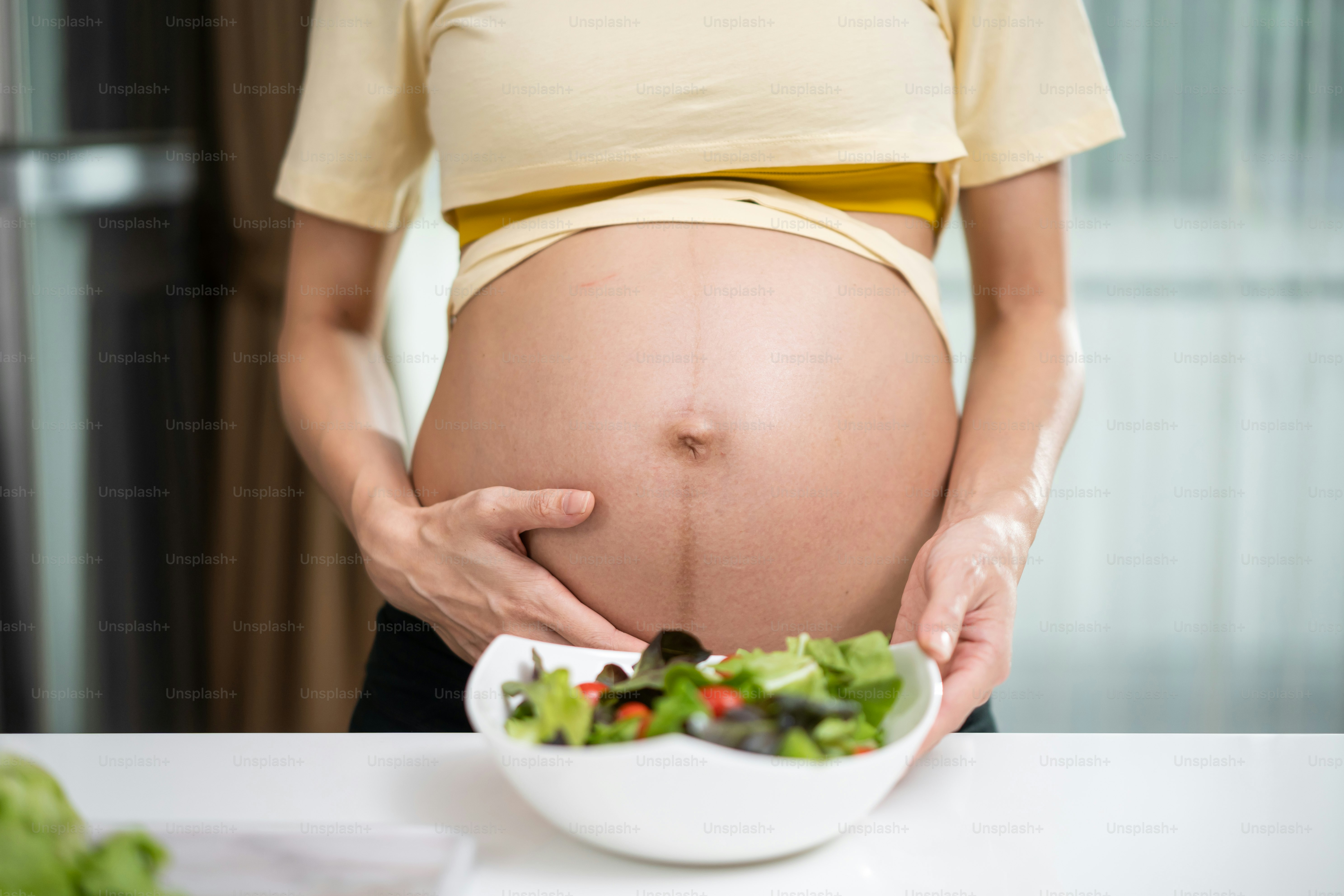 Nahaufnahme einer schwangeren asiatischen Frau, die steht und eine gemischte Schüssel hält. Attraktiver schöner junger weiblicher Schwangerschaftsstand und berührendes Baby im Mutterleib, wählen Sie gesundes Essen oder vegetarisches Essen in der Küche.