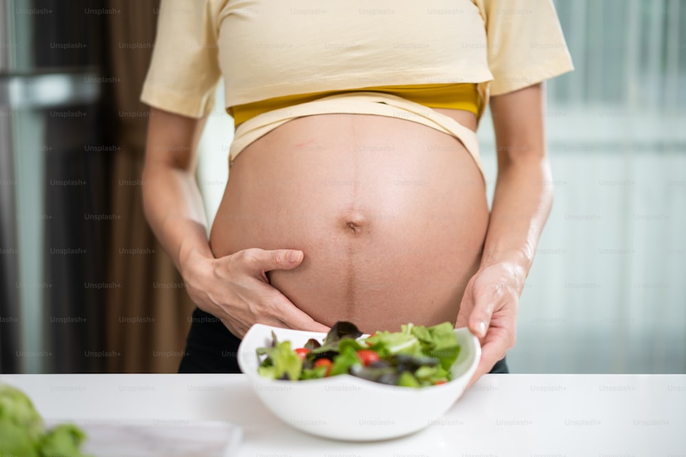 Close up of Asian woman pregnant standing and holding mix slad bowl. Attractive beautiful young female pregnancy stand and touching baby in the womb, choose healthy food or vegetarian food in kitchen.