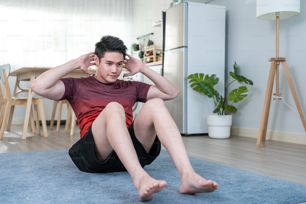 Asian handsome active young man doing sit up on floor in living room. Attractive male in sport cloth spend leisure activity time to exercise and workout on holiday at home. Healthy lifestyle concept.