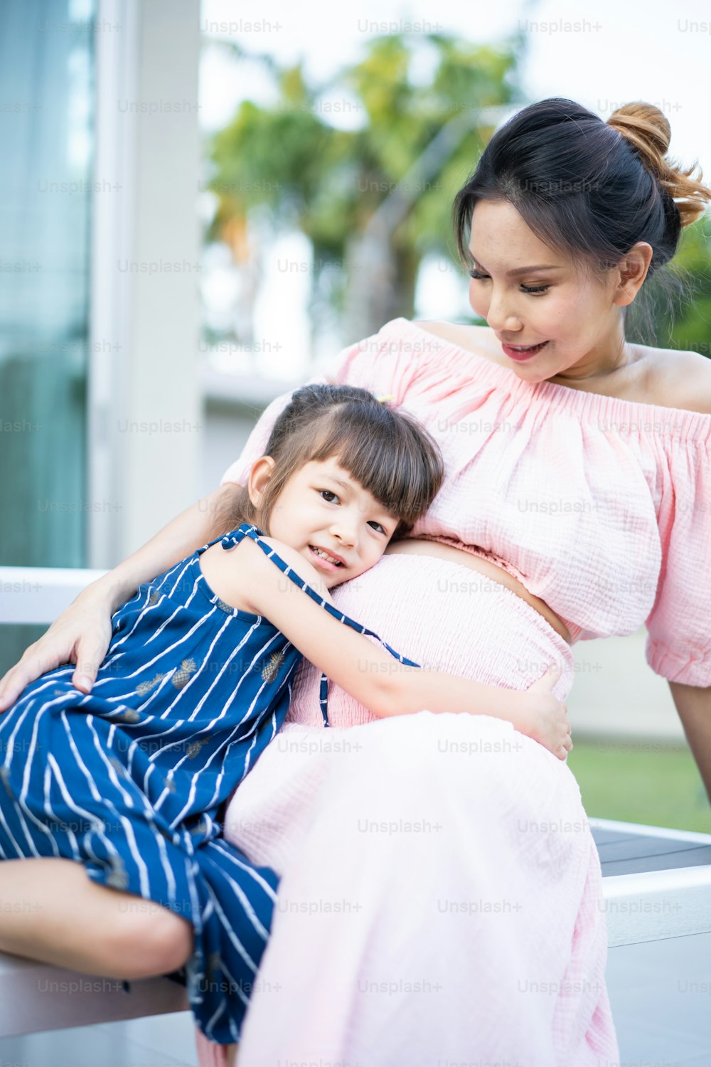 Asian young daughter hug and stroking beautiful pregnant mother tummy. Attractive expecting pregnancy girl embrace little kid girl with happiness and love while stand indoor near window in living room