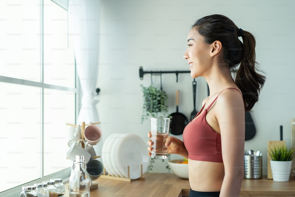 Asian beautiful woman in sportswear drink water after exercise at home. Young thirsty active sport girl takes a sips of clean mineral natural in cup after workout for health care in kitchen in house.
