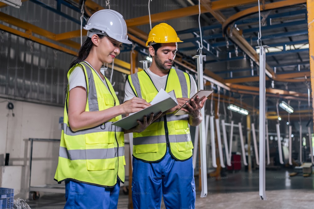 Caucasian male and Asian female industrial worker working in warehouse. Attractive industry factory engineer people wear helmet and processes orders and product at manufacturing plant with happiness.