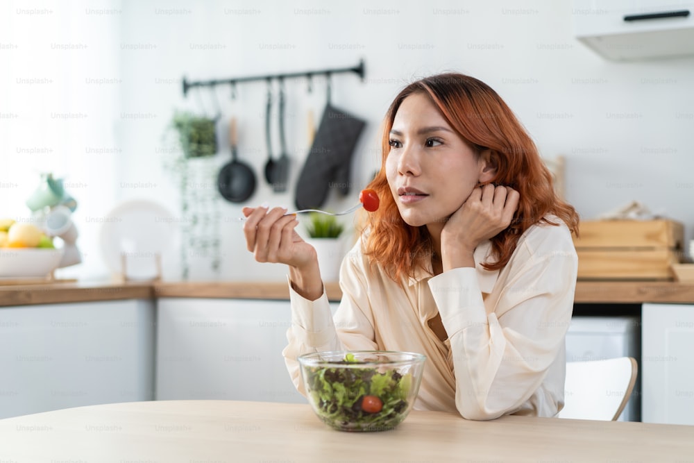 Jovem asiático feliz mulher atraente comer salada verde na cozinha em casa. Menina bonita se sentir alegre e desfrutar de comer vegetais alimentos saudáveis para dieta e perder peso para o bem-estar de cuidados de saúde em casa.