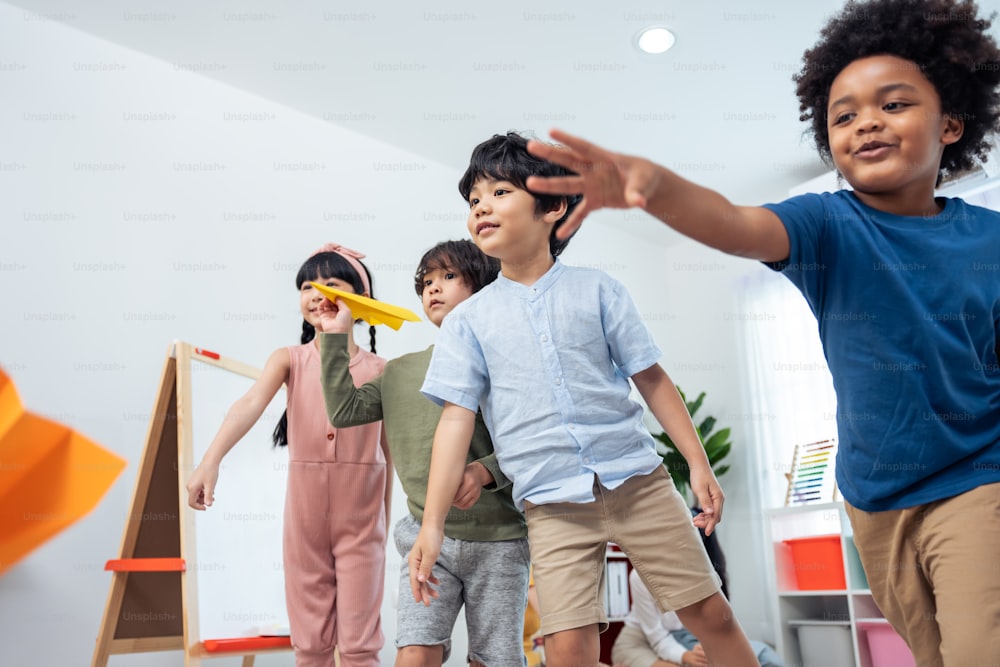 Group of Mixed race young little kid playing airplane in schoolroom. Child girls and boys student spending time play paper rocket while during break with feeling fun together in classroom at school.
