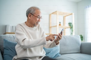Asian senior elderly male using mobile phone in living room at home. Strong older mature grandfather wear eyeglasses, sit on sofa and chat on smartphone communicate with family enjoy retirement life.