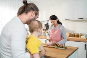 Caucasian beautiful parents cook food with baby boy toddler in kitchen. Happy family, Attractive young mother making healthy salad for lunch while husband father holding little kid son infant in house