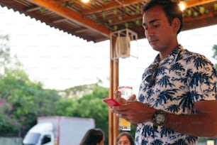 Young man with a beer and a phone in a bar, in the background of a group of friends at a table.