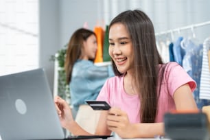 Asian business women enjoy shopping online in web store in warehouse. Young attractive business people work sit on table, hold credit card to make e bank online payment after use labtop purchase goods