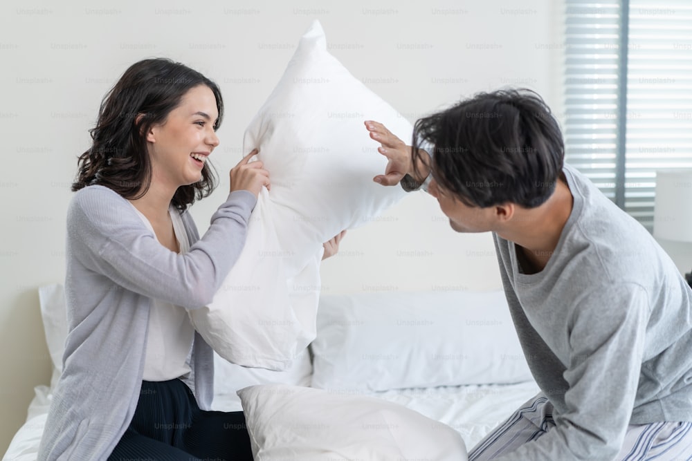 Giovane coppia di innamorati asiatici ha una lotta con i cuscini insieme sul letto. L'uomo e la donna attraenti del nuovo matrimonio si sentono felici, godono dell'attività mattutina che gioca nella camera da letto della casa.