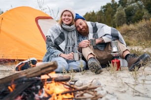 Smiling man holding a cup with tea and looking at the campfire with calmness. Happy family sitting at the nature and drinking hot tea from cups at camp fire in cozy forest