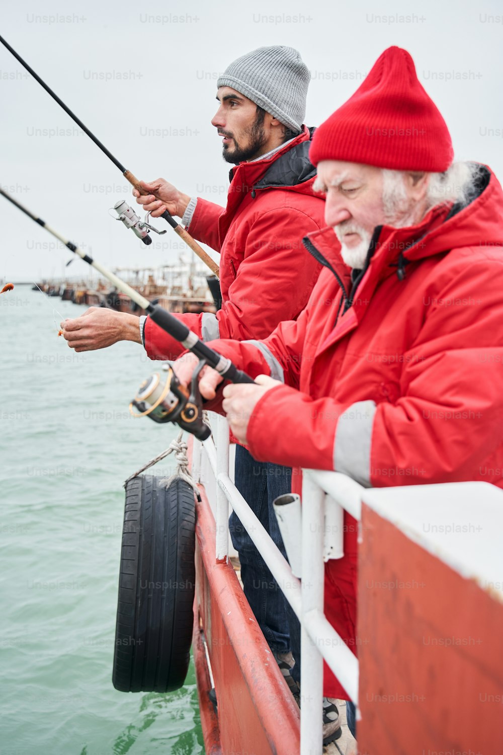 Seitenansicht des glücklichen, selbstbewussten älteren Mannes und seines jungen Kollegen, die bei winterlichem Wetter bei bewölktem Tag unter grauem Himmel auf dem Meer gemeinsam von einem Boot aus fischen. Archivfoto