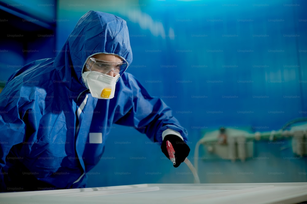 Hombre con ropa de trabajo protectora rociando pintura sobre madera procesada en una instalación de producción.