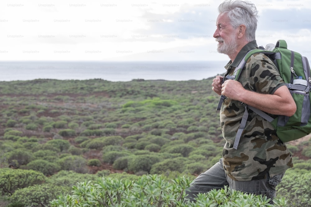 Uomo adulto anziano spensierato che si gode un'escursione all'aperto tra cespugli verdi e mare. Anziani dai capelli bianchi sorridenti con lo zaino