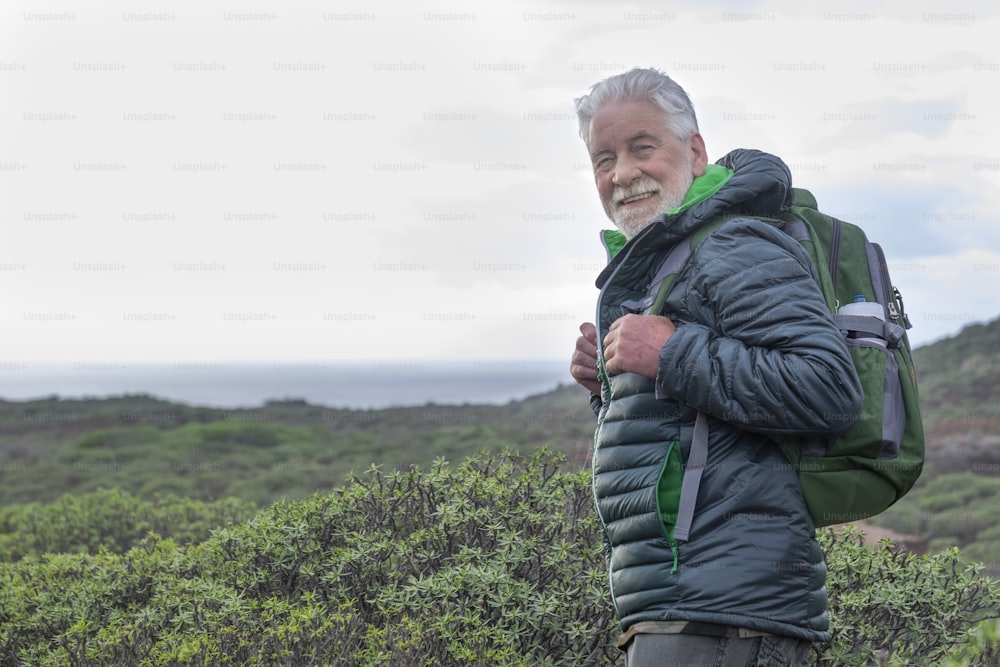 Uomo anziano sorridente dai capelli bianchi che guarda la macchina fotografica durante un'escursione tra cespugli verdi e il mare. Orizzonte sull'acqua. Pensionato attivo zaino in spalla che si gode la vita all'aria aperta e la libertà