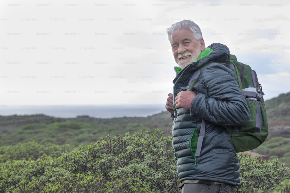 Smiling white-haired elderly man looking at camera while hiking among green bushes and the sea. Horizon over the water. Active retiree backpacker enjoying the outdoors and freedom