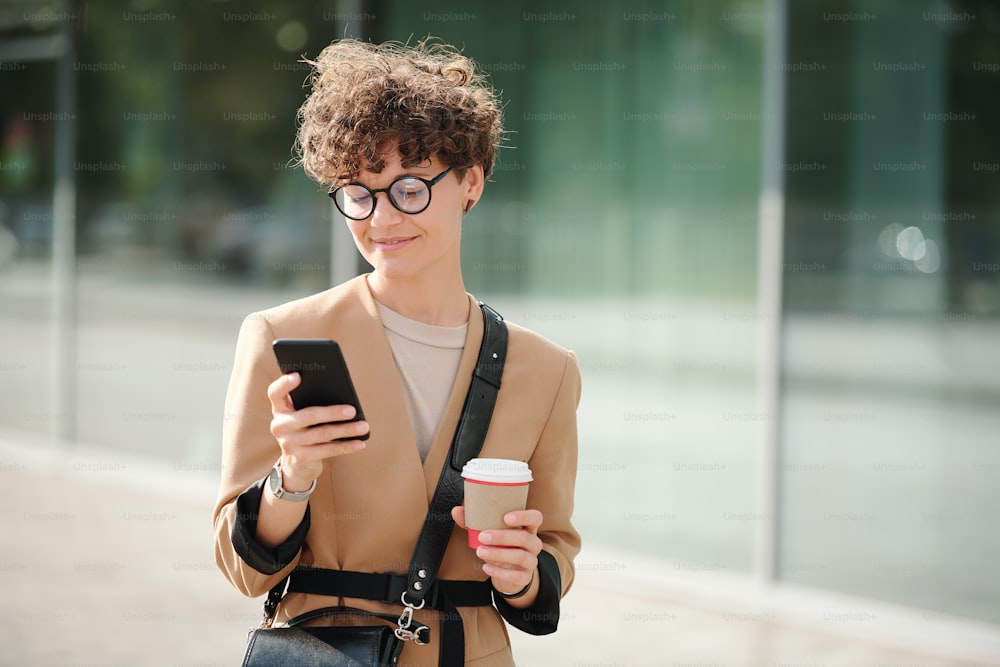 Junge elegante brünette Geschäftsfrau mit Glas Kaffee scrollt durch Kontakte im Smartphone im Freien, während sie telefonieren geht