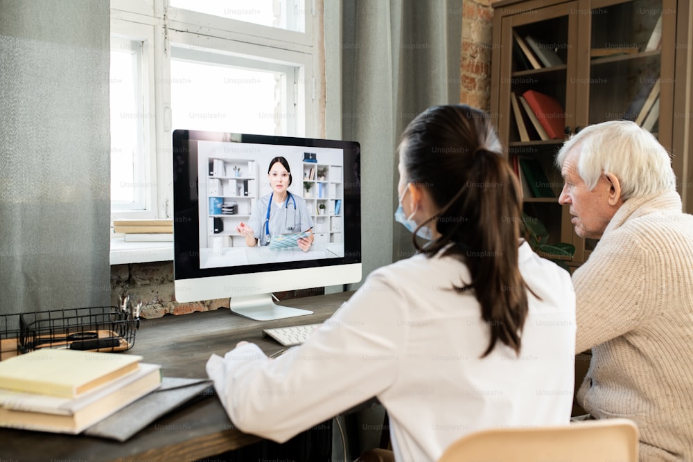 Sick senior man and young female general practitioner sitting in front of computer monitor with doctor on screen giving medical consultation