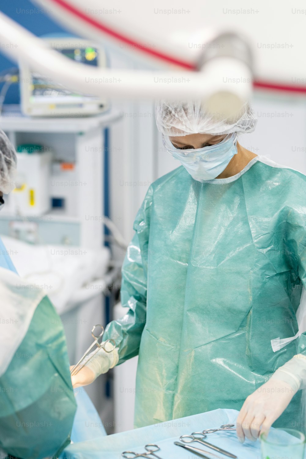 手術中に患者のそばに立ち、医療機器を服用する保護作業服を着た専門外科医の若い女性助手