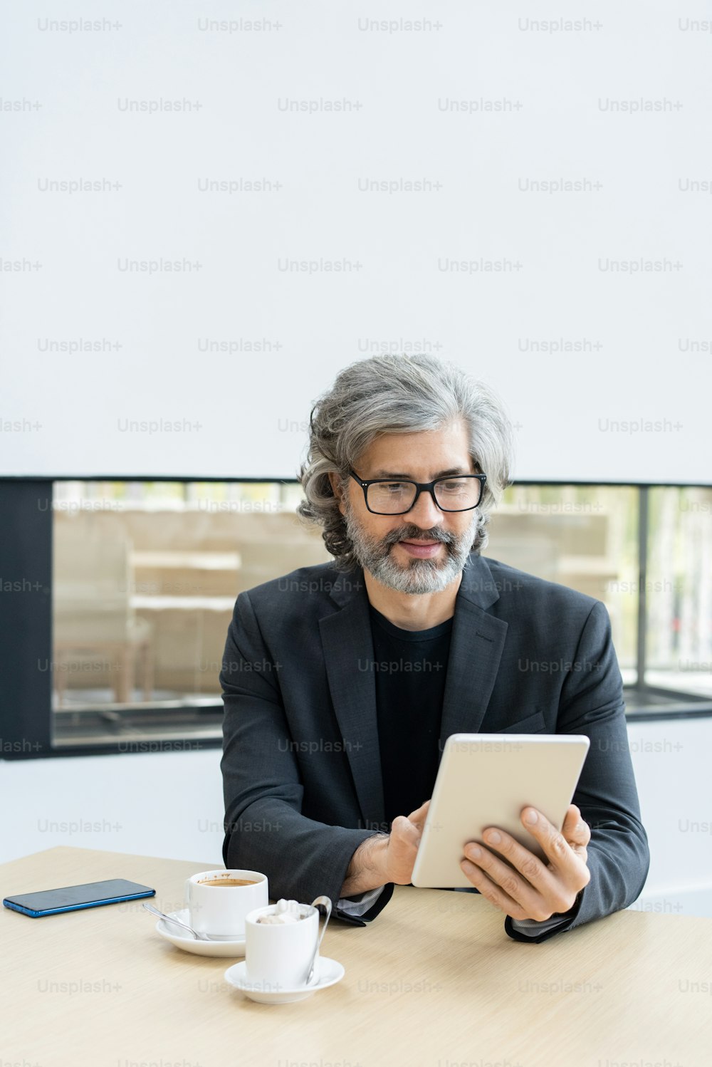 Mature bearded broker with grey hair looking through online news or financial data while sitting by table in cafe of modern business center