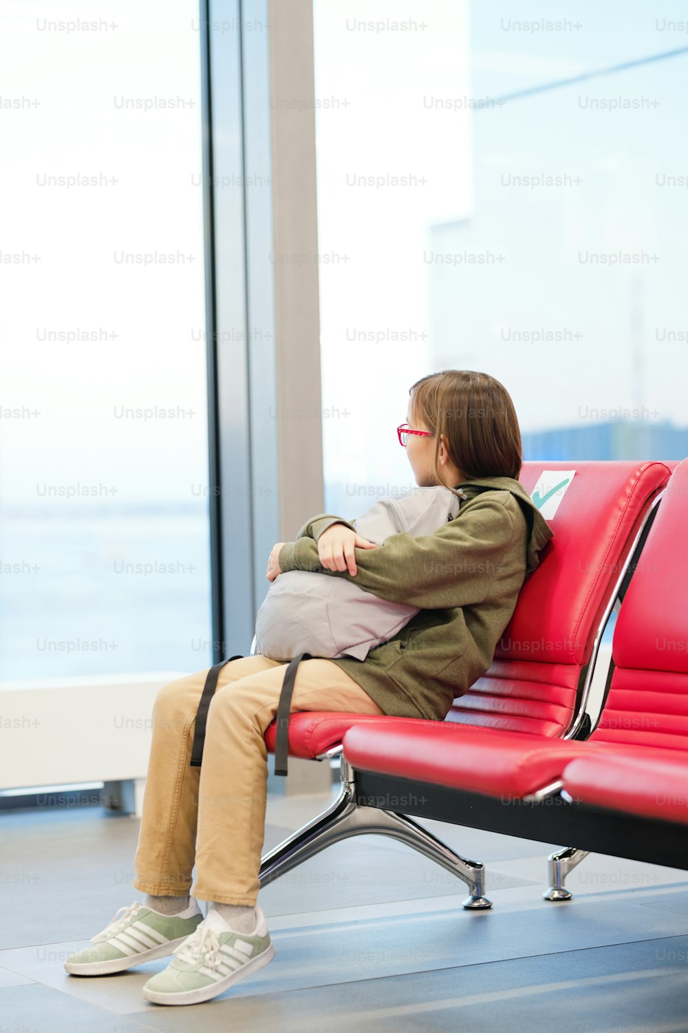 Little girl with backpack sitting on chair and looking at window waiting for her departure in airport terminal