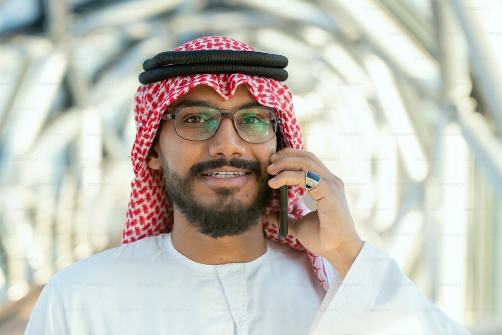 Lächelnder junger arabischer Delegierter oder Geschäftsmann in nationaler Kleidung, der vor laufender Kamera im modernen Geschäftszentrum telefoniert
