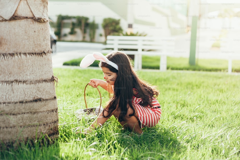 Cute little girl with bunny ears and basket of Easter eggs in the garden. Easter egg hunt