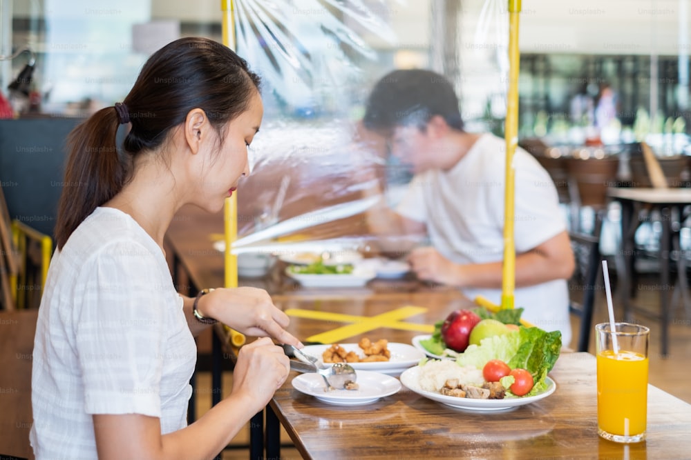 아시아 커플 남녀는 코로나바이러스 covid-19, 레스토랑 및 사회적 거리두기 개념으로부터 감염을 보호하기 위해 테이블 방패로 음식을 먹는 레스토랑에 앉아 있습니다.