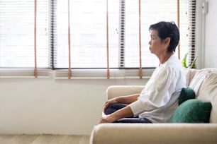 Senior old Asian woman sitting on sofa praticing yoga and meditation at home, retirement and wellbeing concept