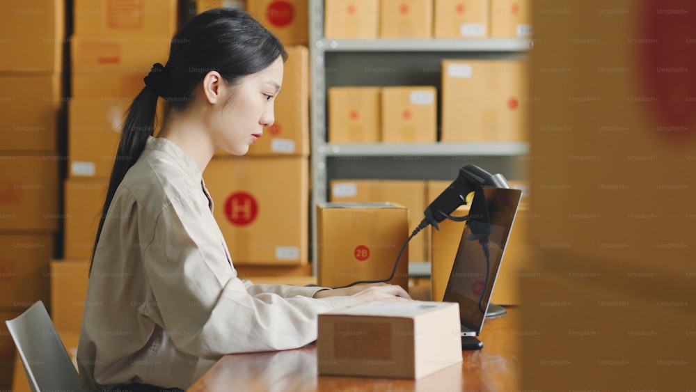 Asian woman working at online store warehouse using laptop computer over parcel boxes on shelves, online e-commerce retail small business concept