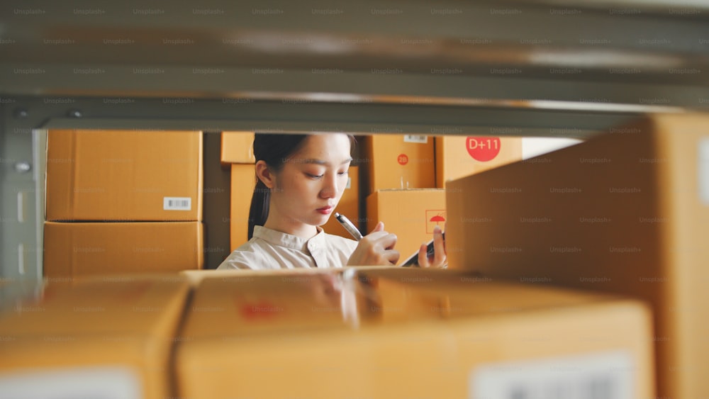 Donna asiatica che lavora al magazzino del negozio online che controlla le scatole dei pacchi di magazzino dell'inventario sugli scaffali, concetto di piccola impresa al dettaglio di e-commerce online