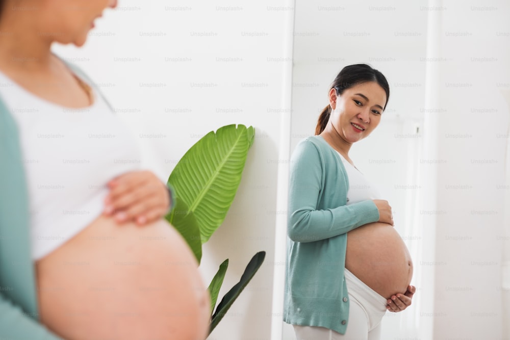 Mujer embarazada asiática que busca espejo con sostén de barriga grande en casa