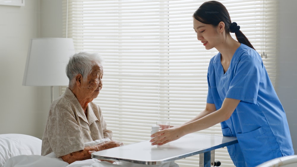 Young Asian woman, nurse, caregiver, carer of nursing home serving medicine pill and glass of water for a senior Asian woman on bed at home.