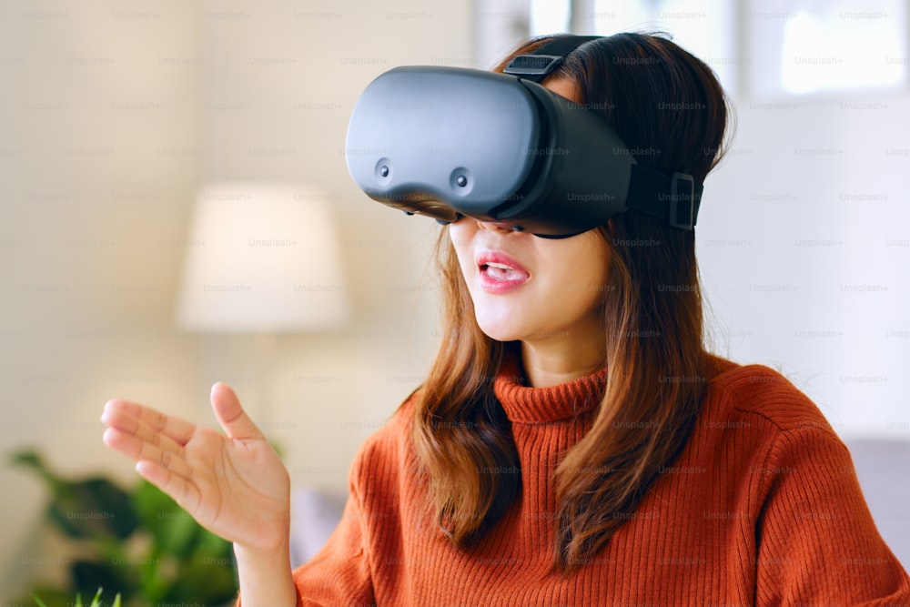 Joven asiática hablando mientras usa auriculares 360 VR para reuniones en línea en el metaverso en casa