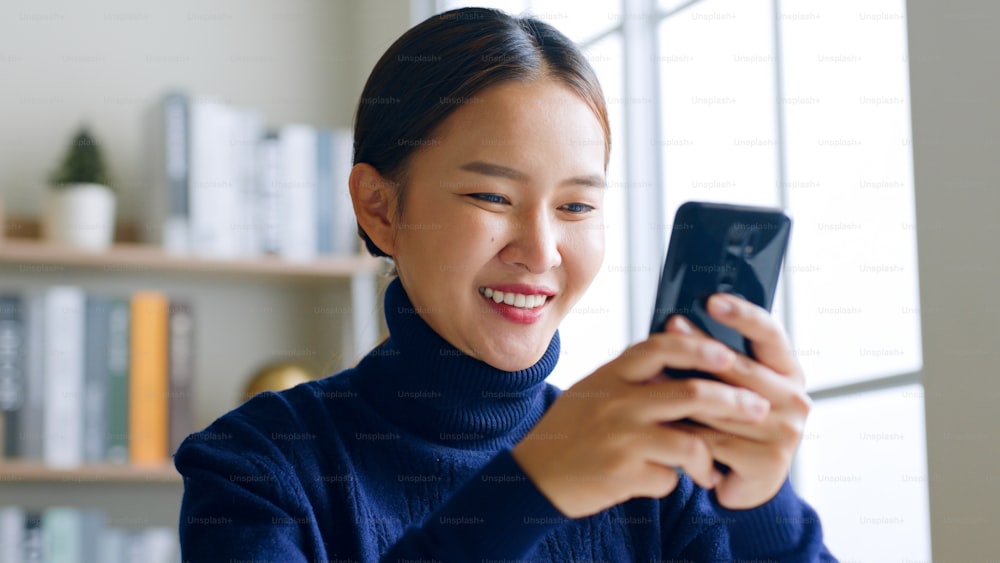 ソーシャルメディアにスマートフォンを使用し、オンラインで買い物をし、家でゲームをして幸せを感じる笑顔の若いアジア人女性
