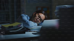Joven empleado de oficina asiático con exceso de trabajo durmiendo mientras trabaja en una computadora portátil horas extras en la oficina por la noche
