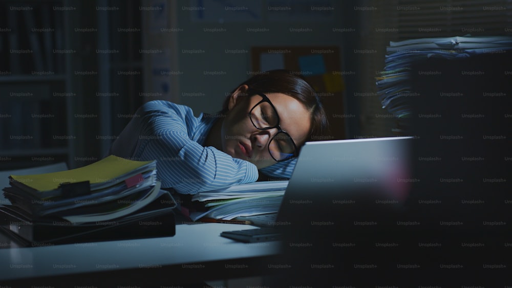 Joven empleado de oficina asiático con exceso de trabajo durmiendo mientras trabaja en una computadora portátil horas extras en la oficina por la noche