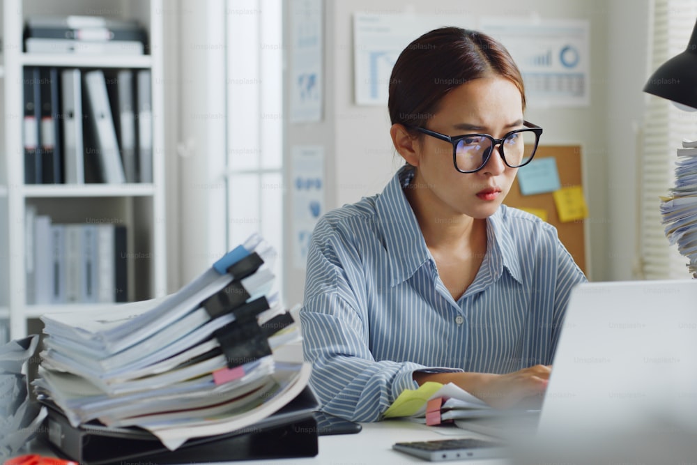 Empregada de escritório asiática que trabalha em laptop, sentindo estresse sério e ocupado no escritório