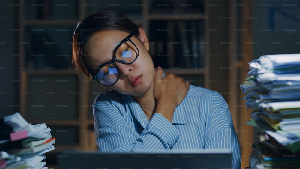 Tired Asian office employee massaging neck and shoulder muscles fatigued from using laptop computer overtime at night in office, office syndrome