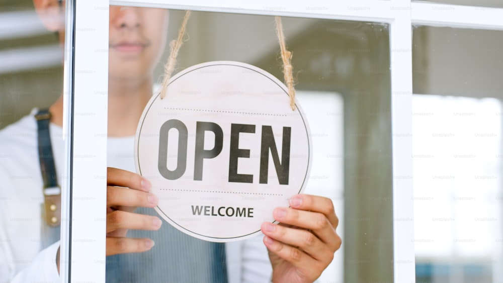 Propietario asiático en delantal que abre el letrero en la puerta de vidrio en la cafetería, tienda, restaurante, concepto de pequeña empresa