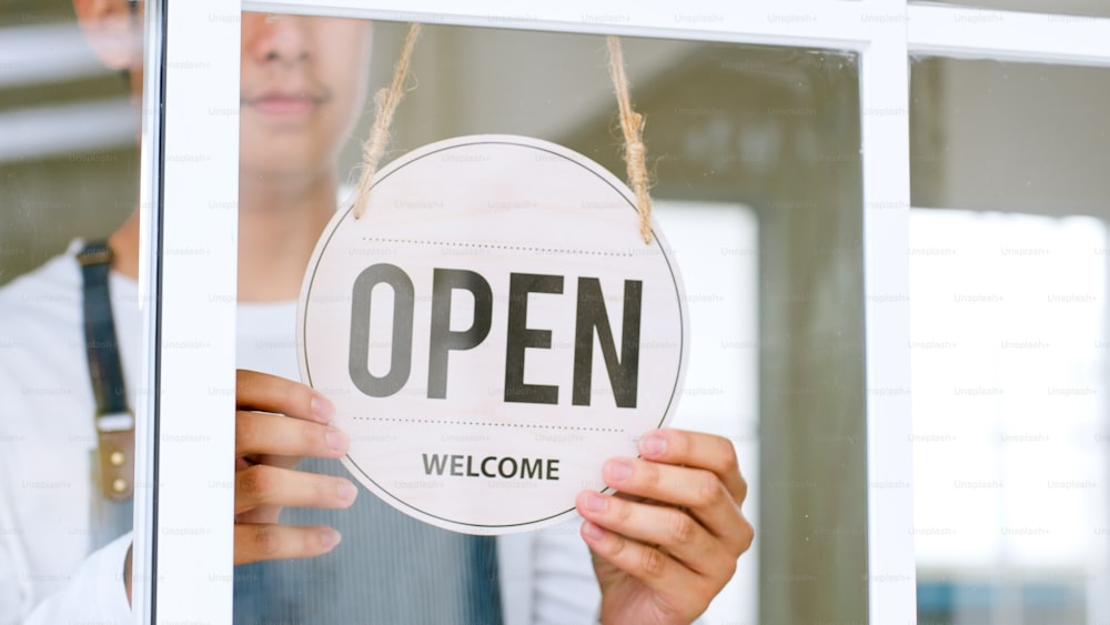 Asian owner in apron turning open sign board on glass door at café, shop, restaurant, small business concept