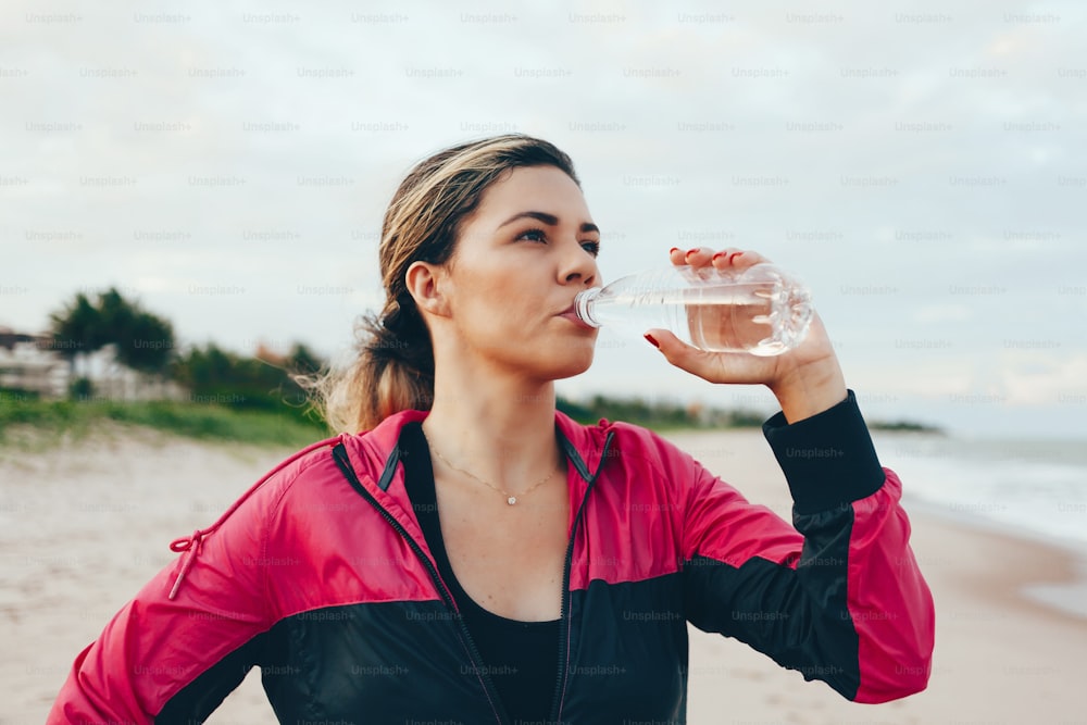 Donna corridore di fitness che beve acqua di una bottiglia sportiva. Ragazza atleta che fa una pausa durante la corsa per idratarsi durante l'esercizio estivo caldo sulla spiaggia. Stile di vita sano e attivo.