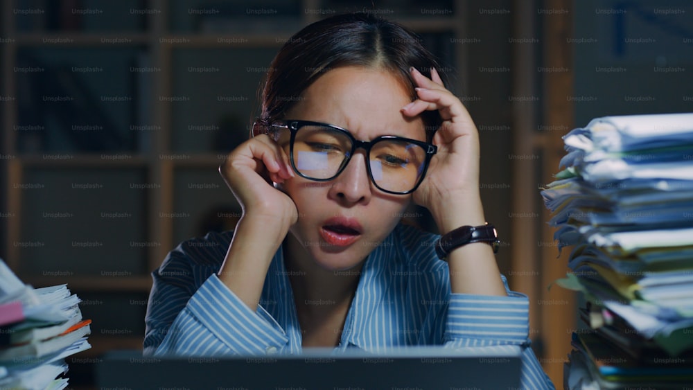 Versuchter asiatischer Büroangestellter, der sich gelangweilt, unglücklich und ausgebrannt fühlt, während er nachts Überstunden im Büro macht