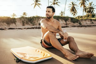 Jovem surfista sentado na praia de areia olhando para o oceano com sua prancha. Conceito de desporto e desporto aquático.