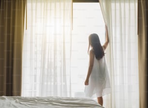 La donna asiatica si sveglia felice al mattino, aprendo le tende sopra la finestra bianca, sentendosi rilassante e fresca
