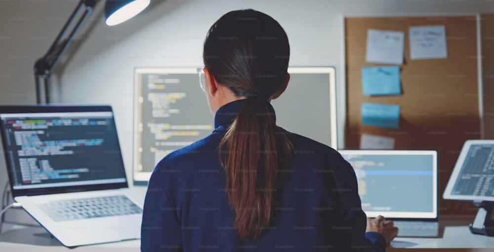 アジアの若い女性、開発者プログラマー、ソフトウェアエンジニア、ITサポート、眼鏡をかけて、盗聴システムのコーディングをチェックするためにコンピューターで残業に一生懸命働いています。背面図