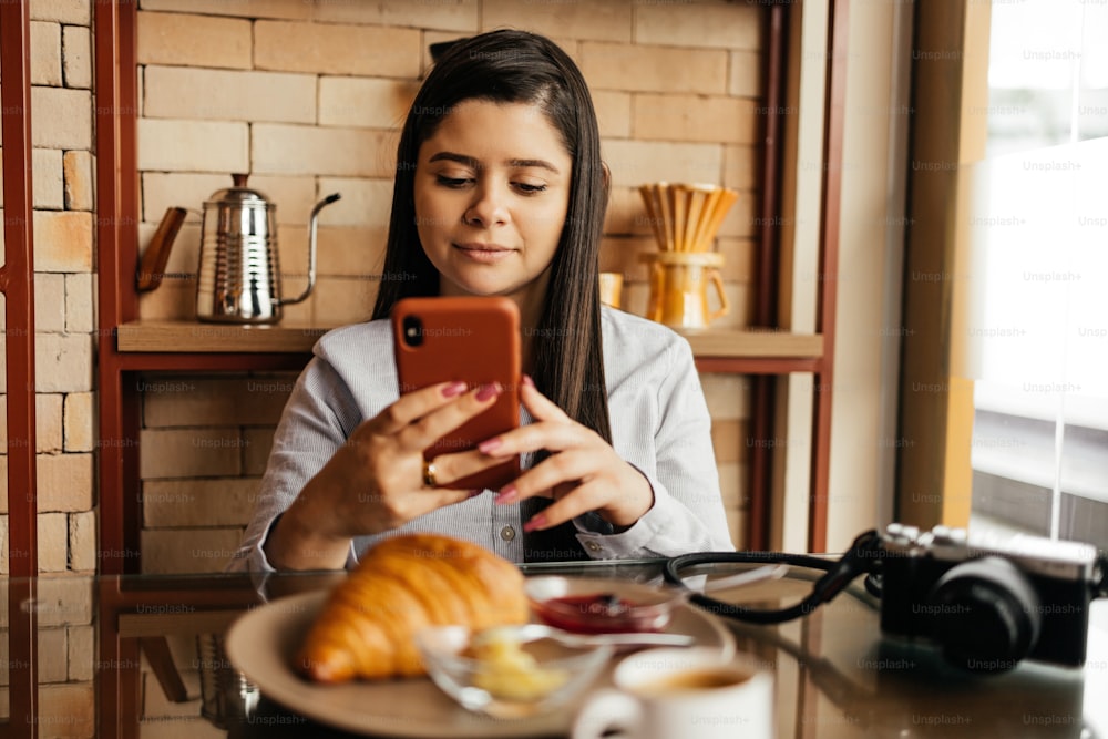 Woman sitting at table and having breakfast with coffee and croissant in cafe while checking emails on cell phone