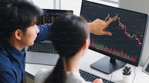 Junge asiatische Investoren, die über Aktienmarkt- und Kryptowährungsinvestitionen aus dem Diagramm im Computer-Laptop zusammen analysieren (verschwommen auf Computer- und Gerätebildschirm)
