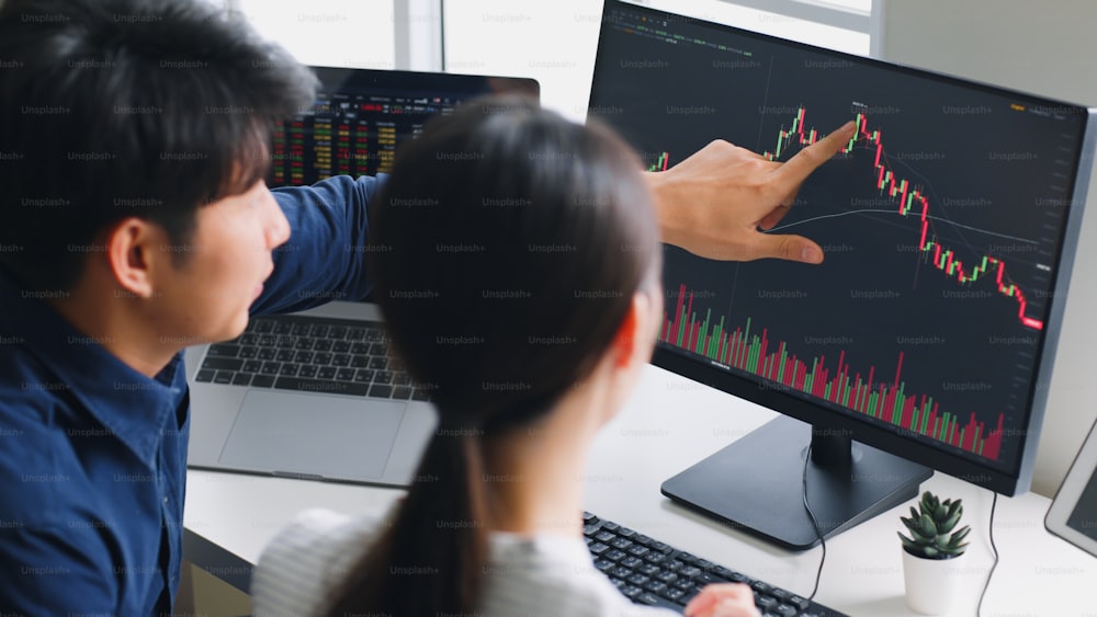 Jovens investidores asiáticos analisando sobre o mercado de ações e o investimento em criptomoedas a partir do gráfico no laptop do computador juntos (borrado na tela do computador e do dispositivo)