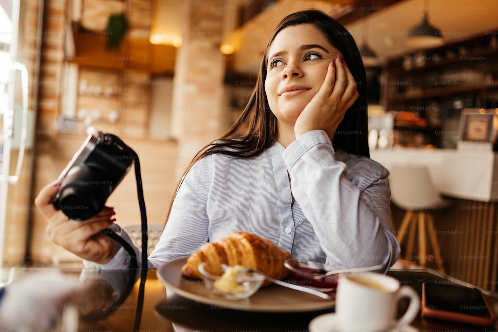 Mulher sonhadora sentada à mesa e tomando café da manhã no café com câmera fotográfica