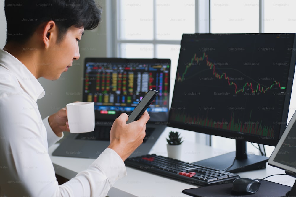 Jeune investisseur asiatique observant l’évolution de la crypto-monnaie et du marché boursier sur smartphone (Flou sur l’écran de l’ordinateur et du mobile)
